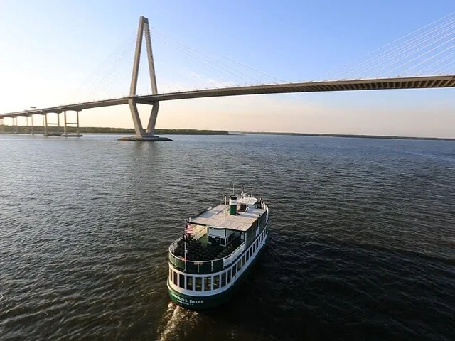 A passenger boat travels on the water toward a large suspension bridge.
