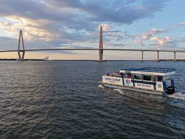 A pontoon boat named Captain Cocktail Cruises sails near a suspension bridge.