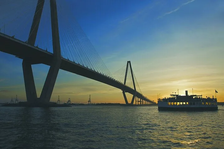 Ein Passagierschiff fährt bei Sonnenuntergang unter einer großen Hängebrücke hindurch.